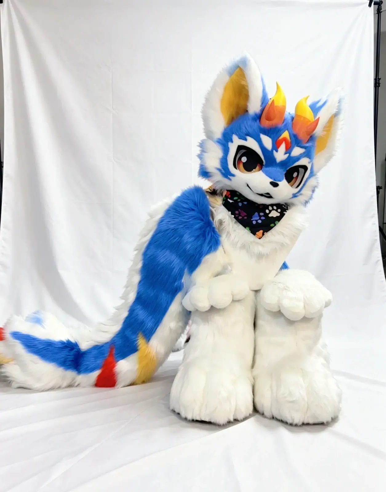 Kemono-style blue and white fursuit with flame-shaped orange horns, amber anime eyes, a paw-print bandana and a long striped tail, seated with the tail curled round