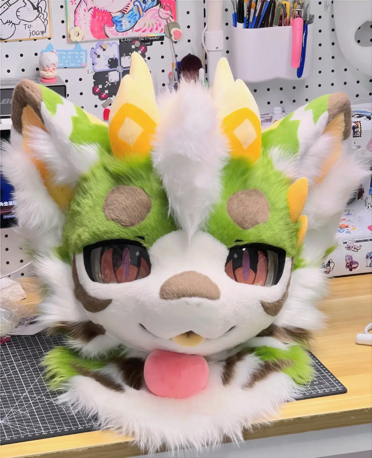 Kemono-style green and white dragon fursuit head with yellow horns, brown spots, large orange anime eyes and a plush pink tongue, front view