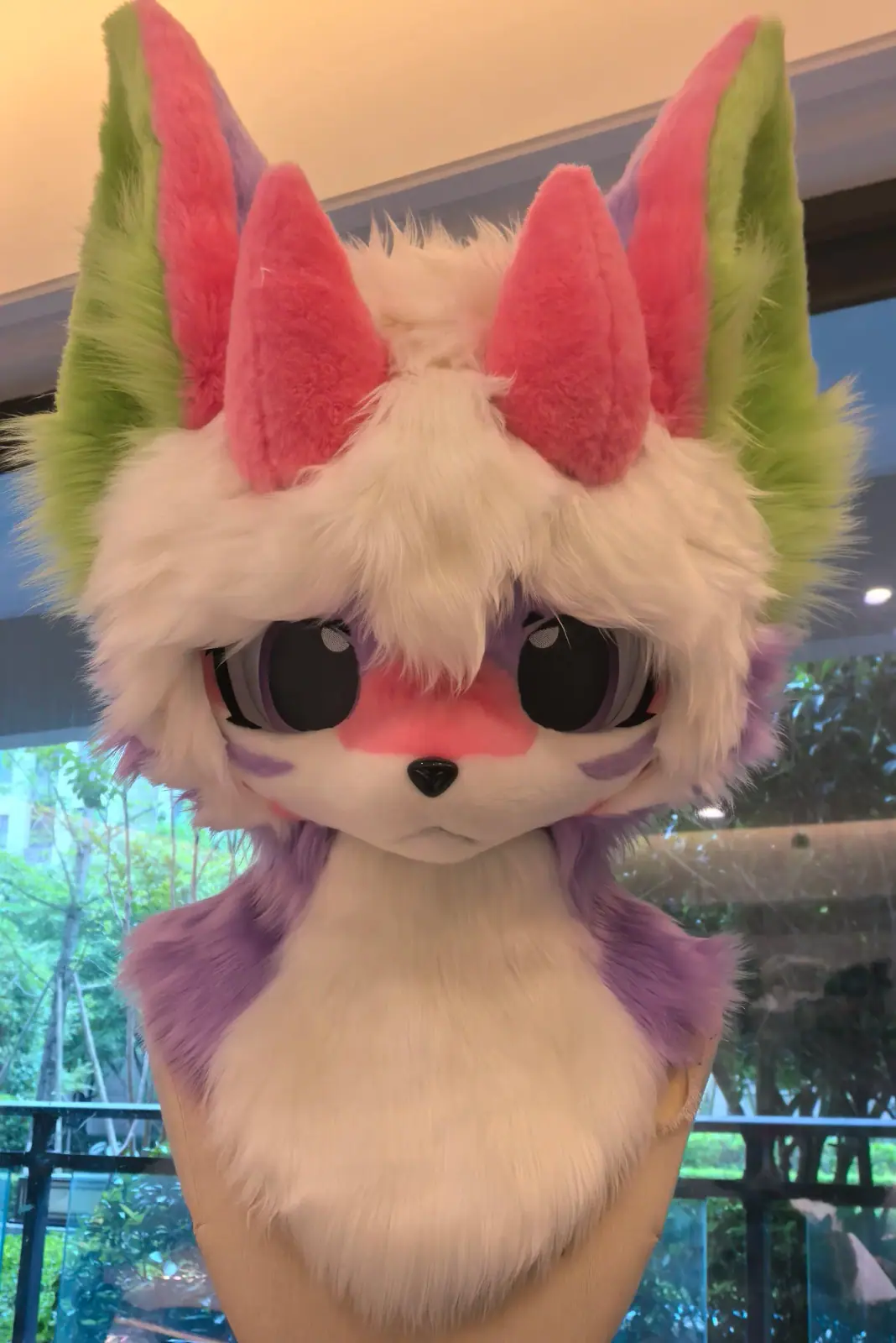 Kemono-style cream fox fursuit head with green, pink and lavender layered ears, pink face markings and big dark anime eyes, front view