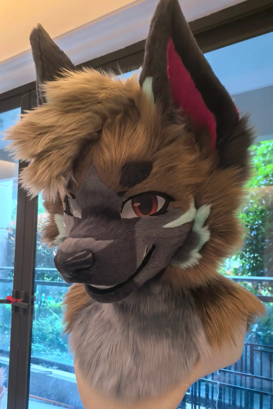 Toony-style brown and grey jackal fursuit head with mint cheek tufts, pink inner ears and a sly smile, three-quarter view in daylight