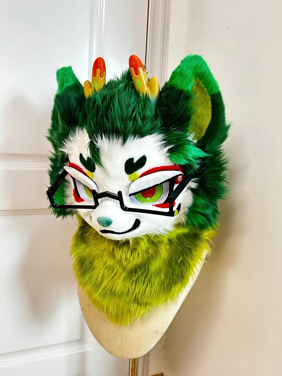 Toony-style white fursuit head with a dark green mane, small orange horns, red-rimmed glasses and a lime chest ruff, front view