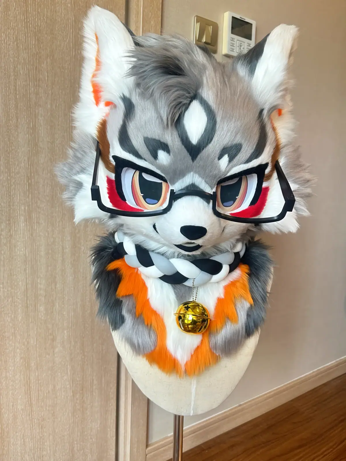 Toony-style grey and white wolf fursuit head with red eye markings, black glasses, a braided collar with a gold bell and an orange chest ruff, front view