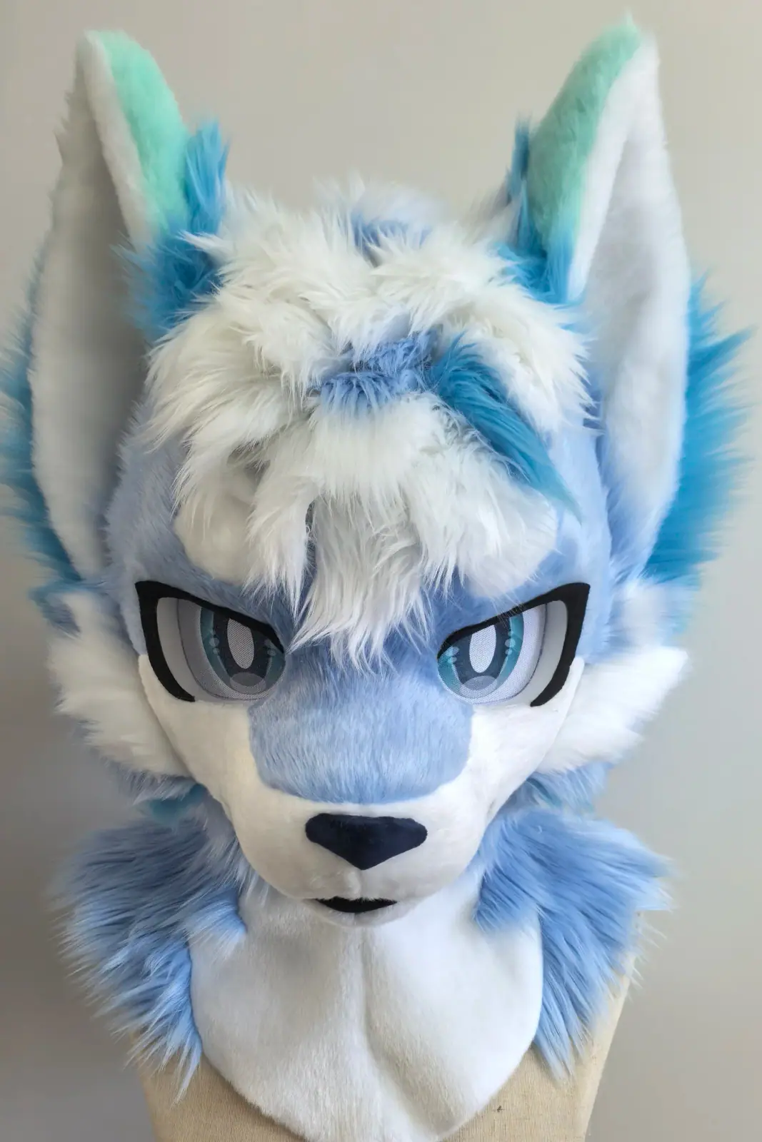 Toony-style ice-blue and white wolf fursuit head with mint ear tips and teal anime eyes, front view