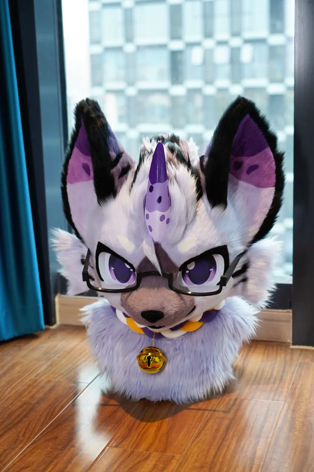 Toony-style lilac and black hyena fursuit head with a spotted purple horn, violet anime eyes, glasses and a bell collar, front view