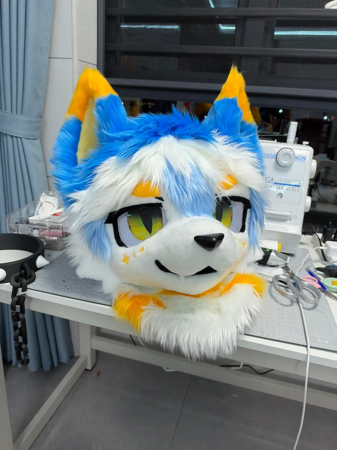 Toony-style white and sky-blue wolf fursuit head with gold ear tips and freckles and big green anime eyes, three-quarter view on the workbench