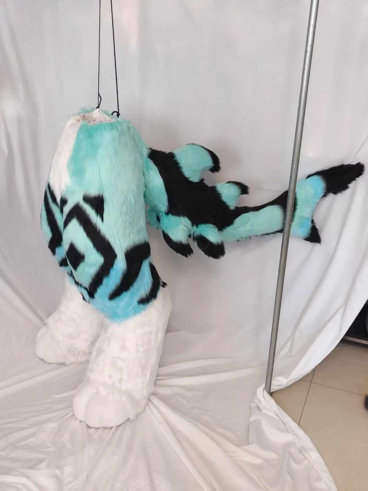 Teal, white and black fursuit bodysuit with a diamond pattern, white feet paws and a finned tail, three-quarter view with the tail