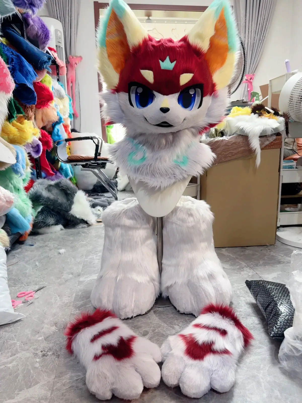 Kemono-style crimson and grey fox fursuit with a teal crown marking, blue anime eyes and red-striped paws, head, hand paws and feet paws in the workshop