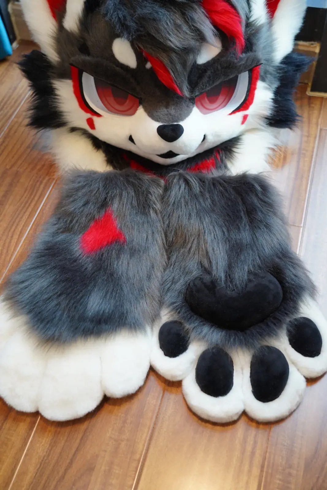 Toony-style charcoal and white wolf fursuit with a red hair streak, red eye markings, red inner ears and matching feet paws, with the matching feet paws